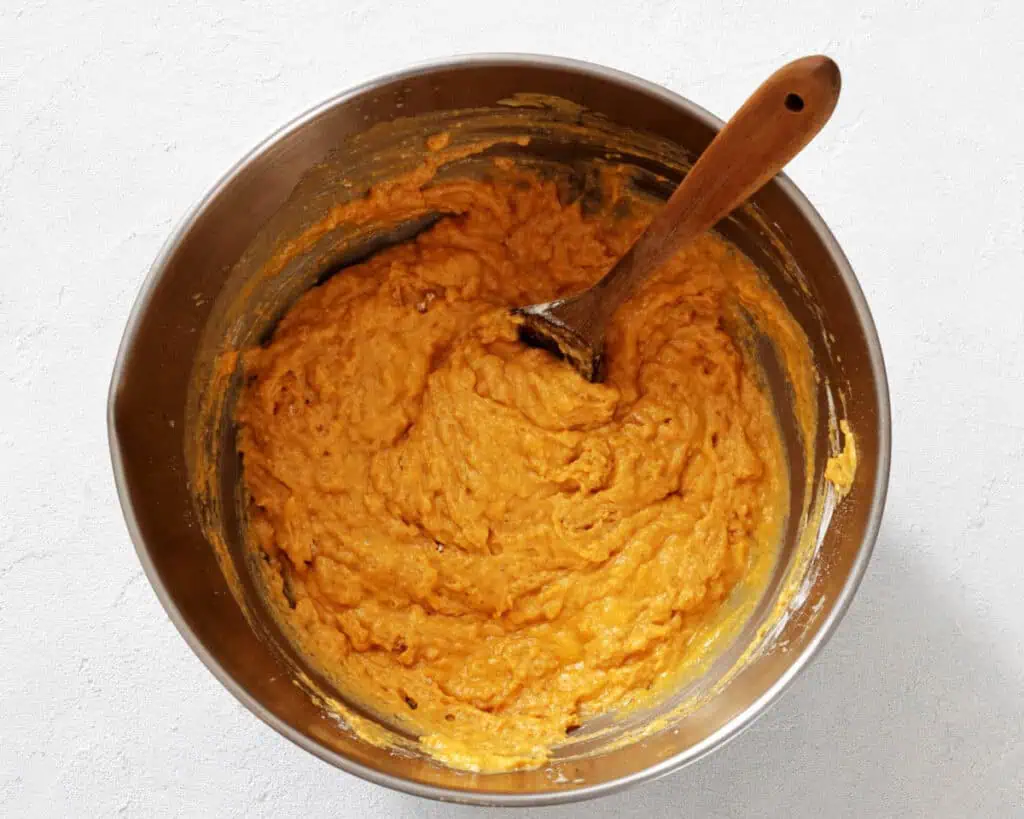 A metal mixing bowl filled with orange batter and a wooden spoon resting inside, placed on a light-colored surface.