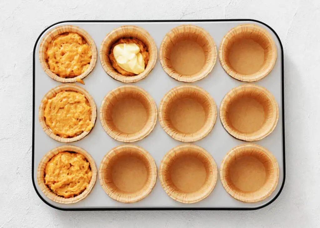 A muffin tin holds twelve paper liners, with three partially filled with batter and one containing a dollop of butter; the rest are empty.