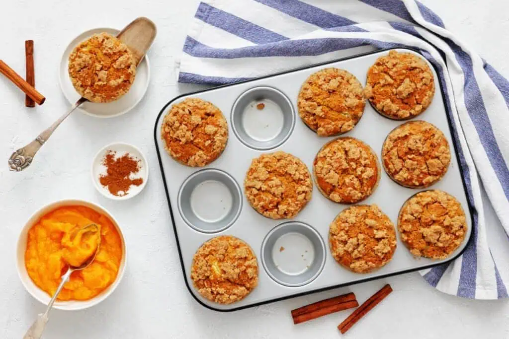 A muffin tin holds nine pumpkin muffins with crumb topping, with three empty spaces. Nearby are a bowl of pumpkin puree, a bowl of spices, a spoon, and a striped towel.