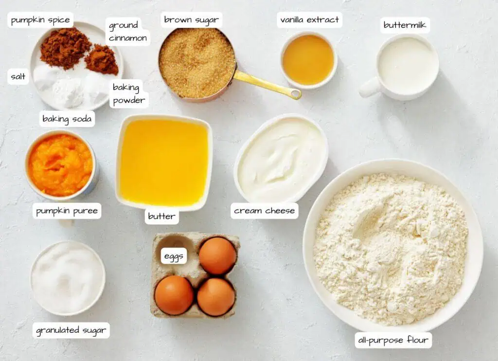 Assorted baking ingredients including flour, eggs, sugars, pumpkin puree, butter, cream cheese, spices, leavening agents, vanilla extract, and buttermilk arranged on a white surface.