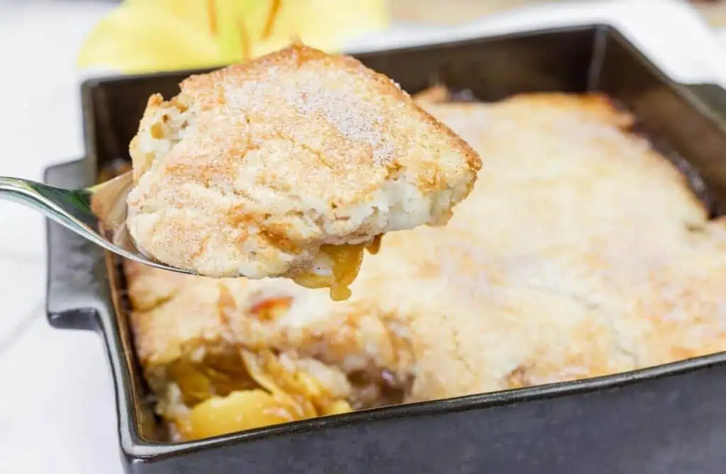 A close-up of a spoon holding a serving of peach cobbler above a baking dish filled with the dessert.