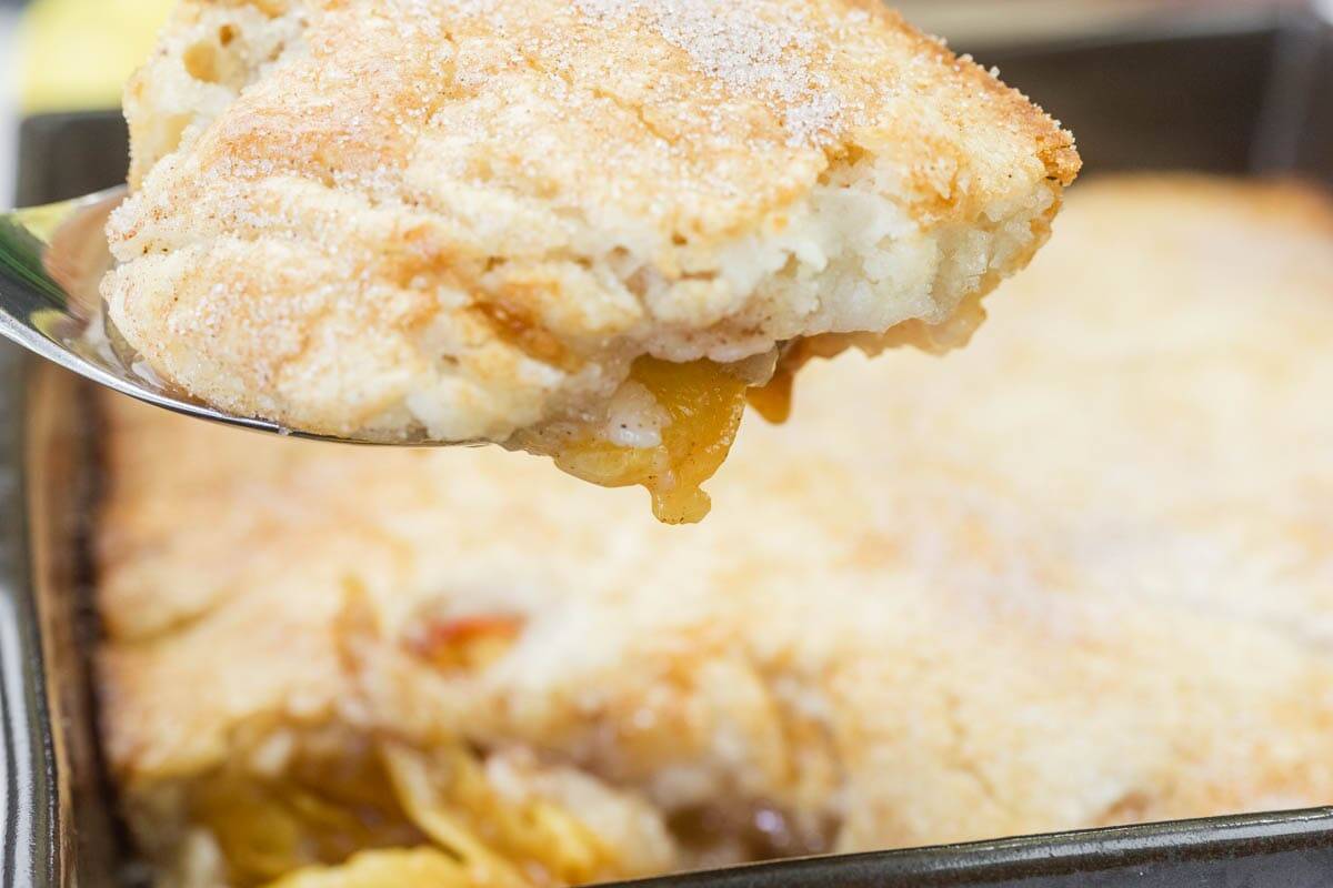A close-up of a serving of peach cobbler being lifted from a baking dish, showing a golden, flaky crust with visible peach filling.