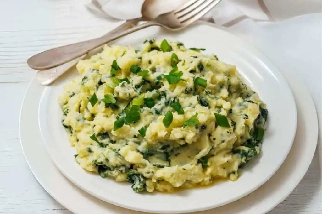 A plate of mashed potatoes mixed with greens, garnished with chopped scallions, served on a white plate with a fork and spoon in the background.