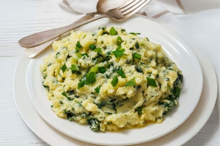 A plate of mashed potatoes mixed with greens, garnished with chopped scallions, served on a white plate with a fork and spoon in the background.