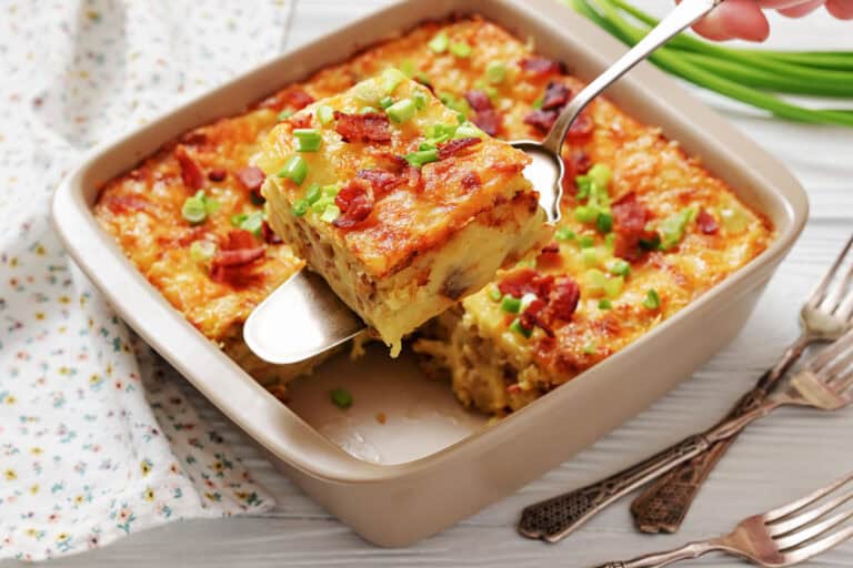A baked casserole topped with chopped green onions and bacon, with one square piece being lifted out on a spatula. Three forks are placed beside the dish.
