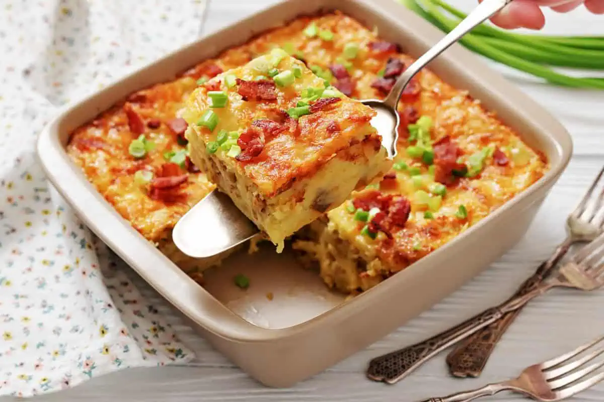 A baked casserole topped with chopped green onions and bacon pieces, with one square being served from the dish using a spatula.