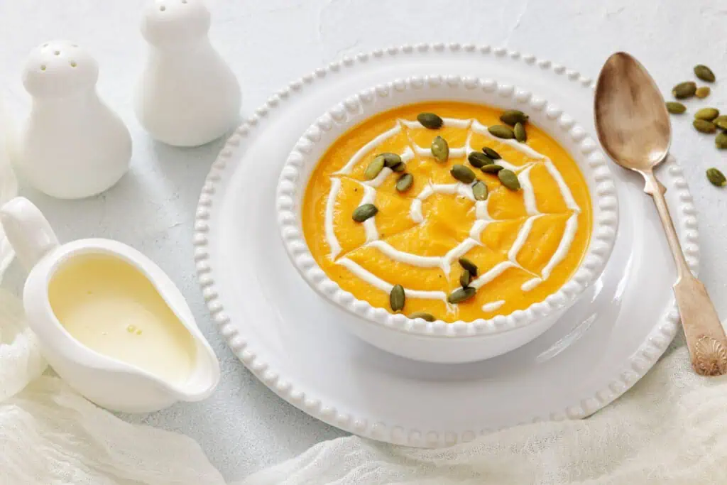 A bowl of creamy pumpkin soup topped with cream and pumpkin seeds, served on a white plate with a spoon, salt and pepper shakers, and a small jug of cream on the side.