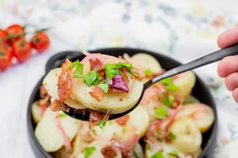 A close-up of a spoon lifting sliced potatoes topped with bacon, red onions, and fresh parsley from a bowl.