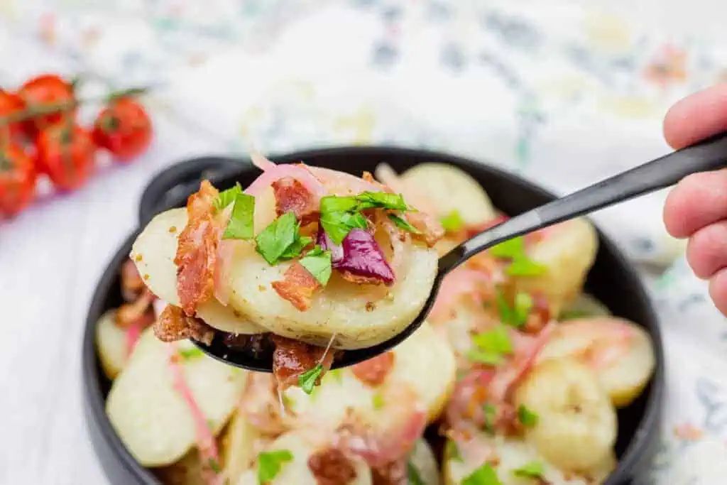 A spoonful of German potato salad topped with bacon, red onion, and chopped parsley held over a bowl of the same salad.