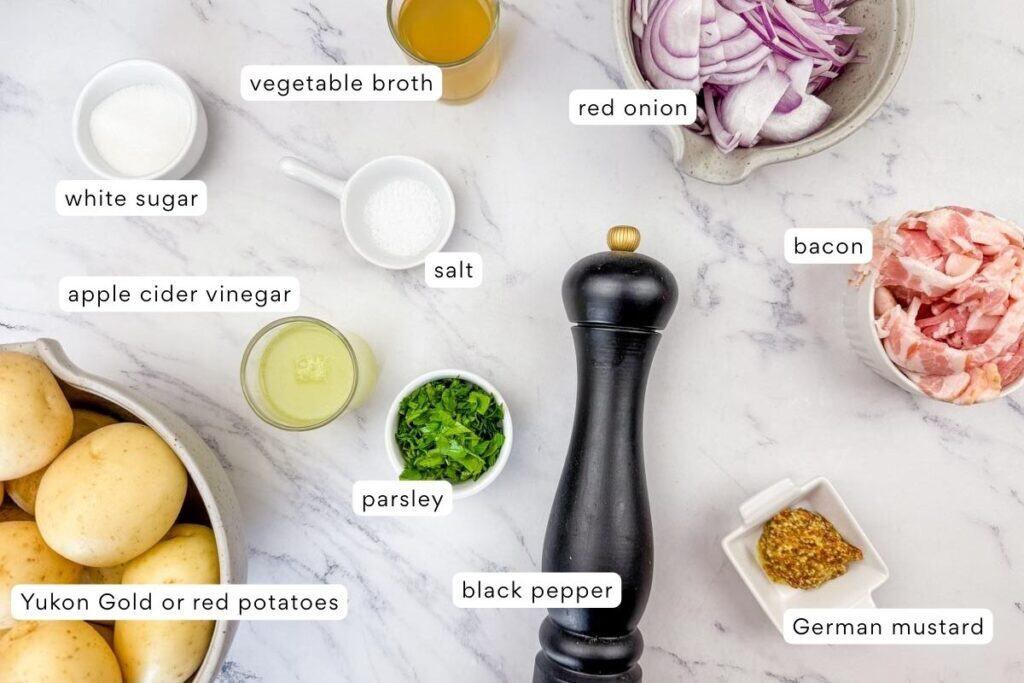 Ingredients for German potato salad are arranged on a marble surface, including potatoes, bacon, red onion, parsley, mustard, vinegar, vegetable broth, white sugar, salt, and black pepper.