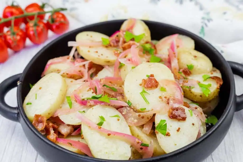 A black dish filled with sliced potatoes, red onions, bacon pieces, chopped parsley, and mustard seeds. Cherry tomatoes are in the background.
