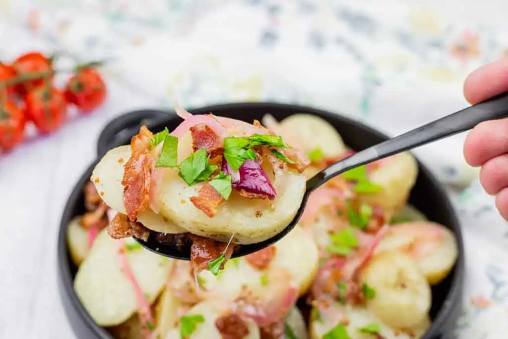 A spoonful of sliced potato salad with bacon, onions, and chopped parsley, held above a bowl of the same dish.