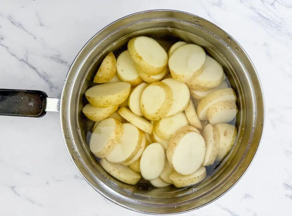A stainless steel pot filled with sliced potatoes submerged in water, placed on a white marble surface.
