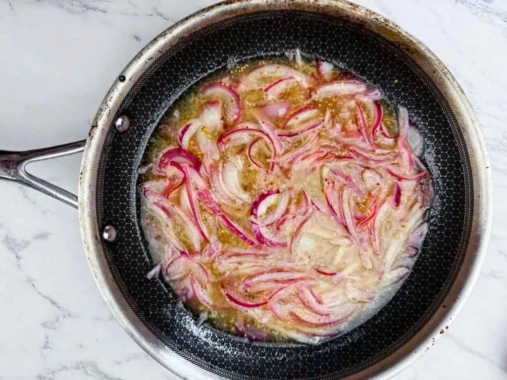 A frying pan on a marble surface containing sliced red onions cooking in oil or liquid.