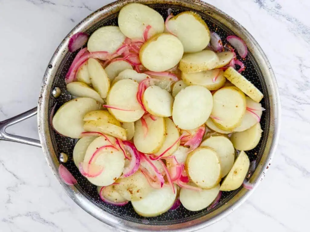 Sliced potatoes and red onions cooking in a stainless steel skillet on a white marble surface.
