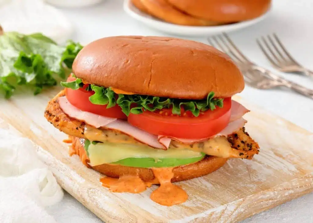 A chicken sandwich with lettuce, tomato, ham, cheese, avocado, and sauce on a bun, placed on a wooden board.