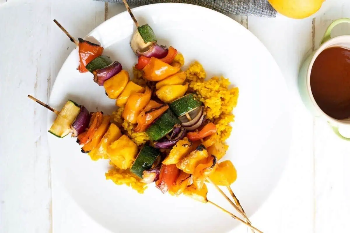 Three Grilled Teriyaki Mango Skewers with bell peppers, zucchini, and onions served on a bed of yellow rice on a white plate.