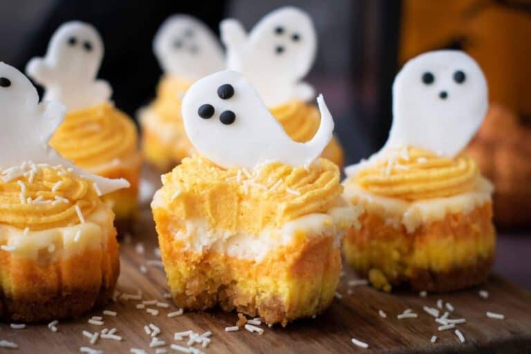 Mini cheesecakes with orange frosting and ghost-shaped fondant toppers are displayed on a wooden surface, sprinkled with white shredded coconut.