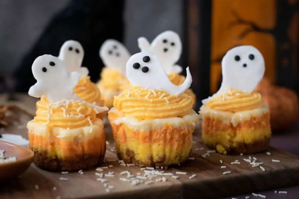 Mini cupcakes with orange and yellow frosting are topped with small ghost-shaped decorations and white sprinkles, arranged on a wooden board.