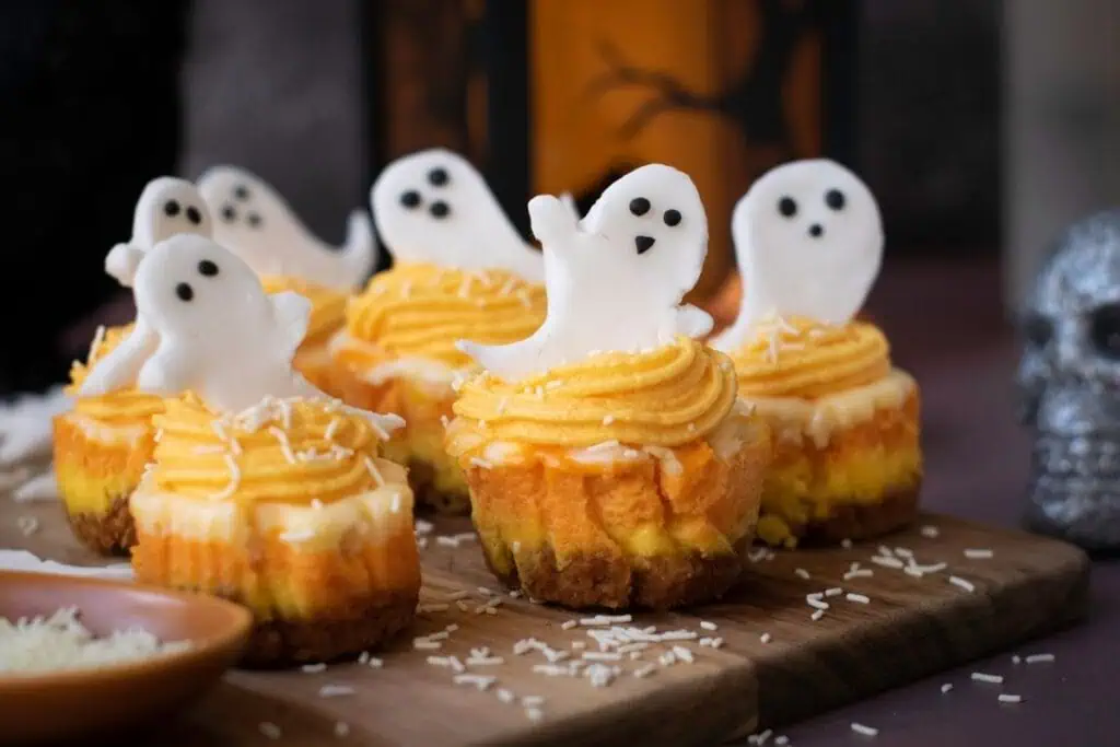 Cupcakes with yellow frosting and white fondant ghost decorations are arranged on a wooden board, with shredded coconut sprinkled on top.