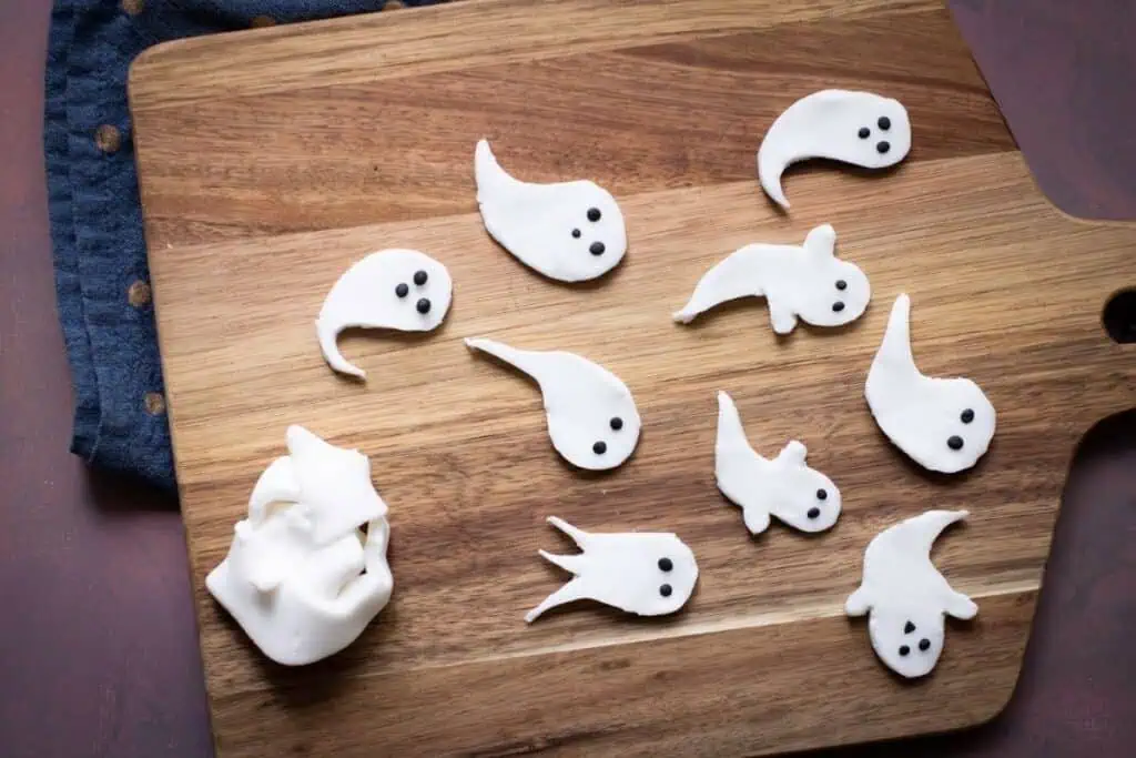 A wooden cutting board with ghost-shaped pieces of white dough, each with two black dots for eyes, and one ball of dough on the left.