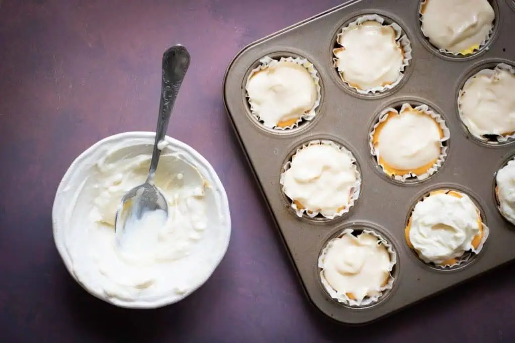 A muffin tin with cupcakes topped with frosting next to a bowl of frosting and a spoon on a dark surface.