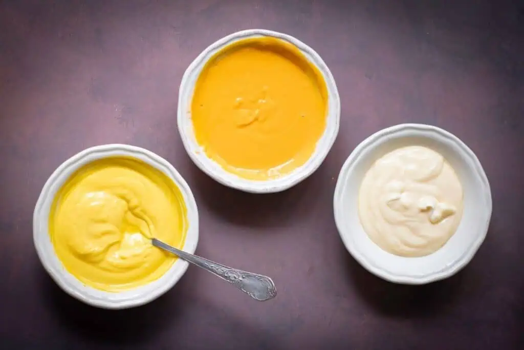Three white bowls filled with different creamy sauces—yellow, orange, and off-white—are arranged on a dark surface, with a spoon in the yellow sauce.