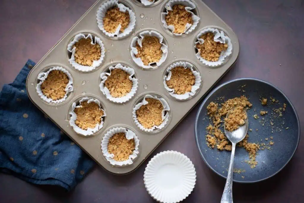 A muffin tin with paper liners holds crushed graham cracker crusts. A plate with crumbs and a spoon is beside the tin, and a blue cloth is partly visible.