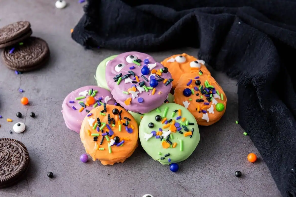 A group of Halloween Oreo Cookies coated in green, orange, and purple, decorated with sprinkles and candy eyes, sits on a gray surface next to a black cloth.