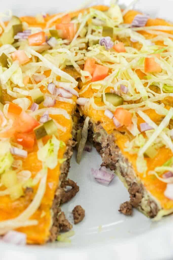 A close-up of a cheeseburger pie topped with shredded lettuce, diced tomatoes, pickles, and chopped onions, with a slice missing.