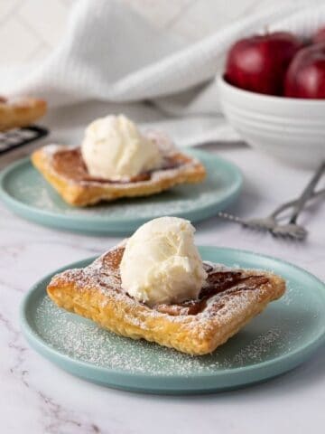Two square apple pastries topped with vanilla ice cream are served on blue plates, with a bowl of red apples and a cooling rack with more pastries in the background.