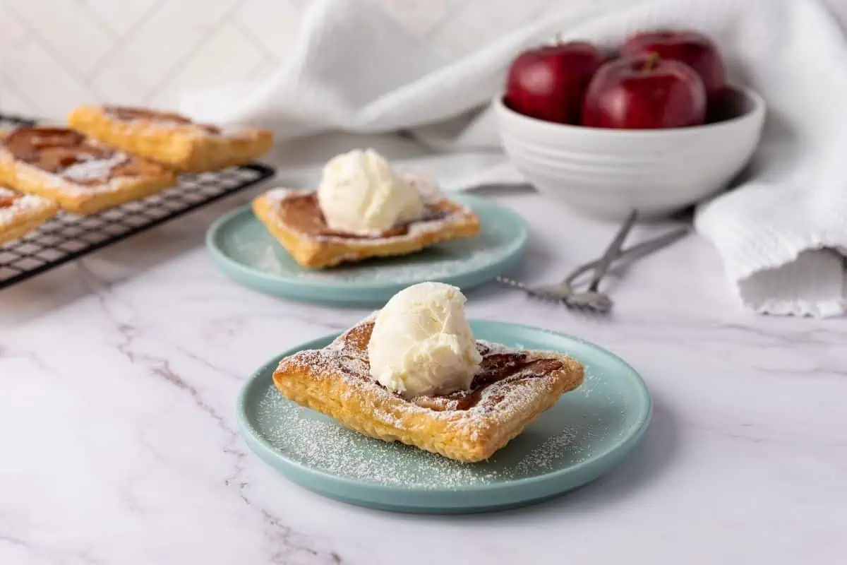 Two square apple pastries topped with vanilla ice cream are served on blue plates, with a bowl of red apples and a cooling rack with more pastries in the background.