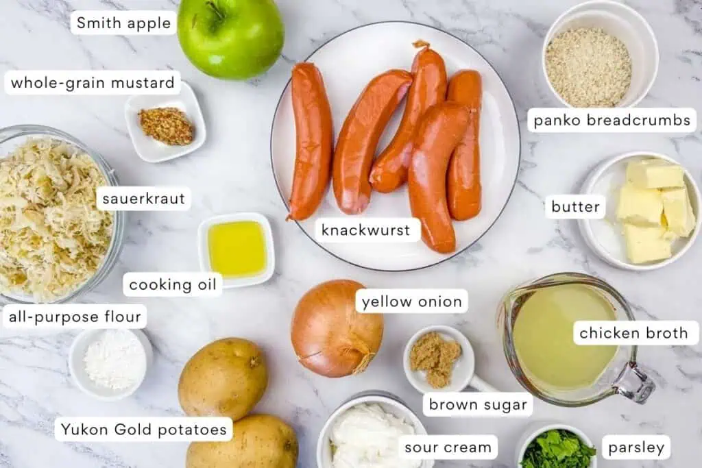 A variety of labeled ingredients on a marble surface, including knackwurst, potato, apple, breadcrumbs, sauerkraut, mustard, onion, butter, and various seasonings.