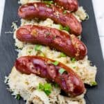 Four grilled sausages are served on a bed of sauerkraut and garnished with chopped herbs on a black rectangular plate.