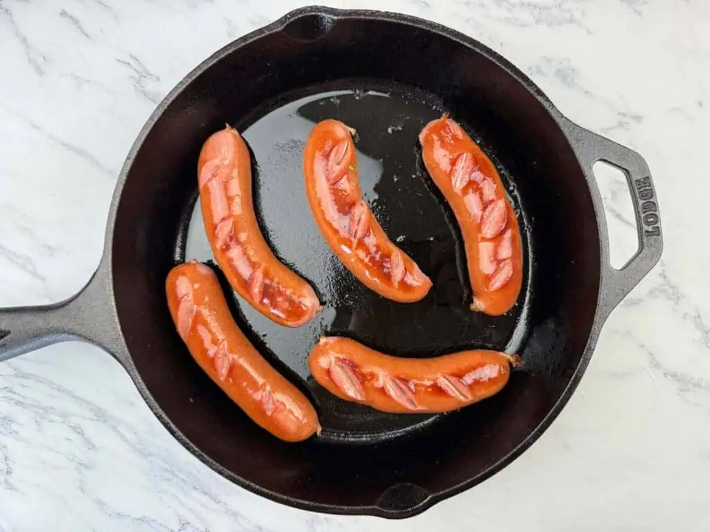 Five scored hot dogs are cooking in a black cast iron skillet on a white marble surface.