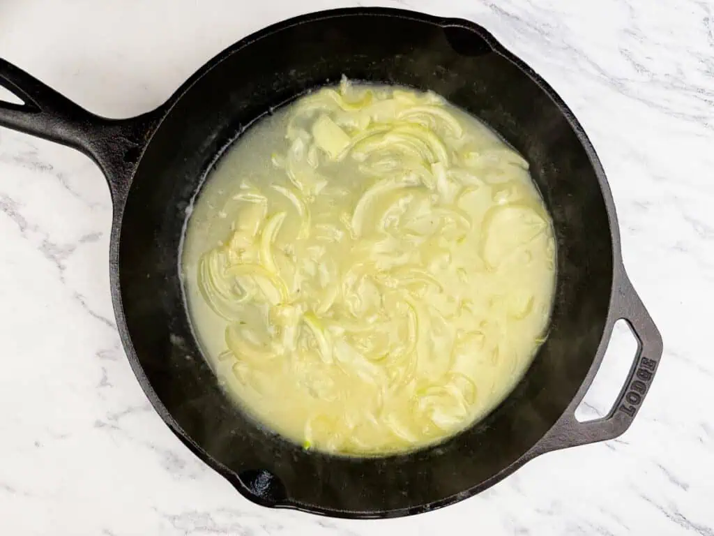 Sliced onions cooking in a creamy sauce inside a black cast iron skillet on a white marble countertop.