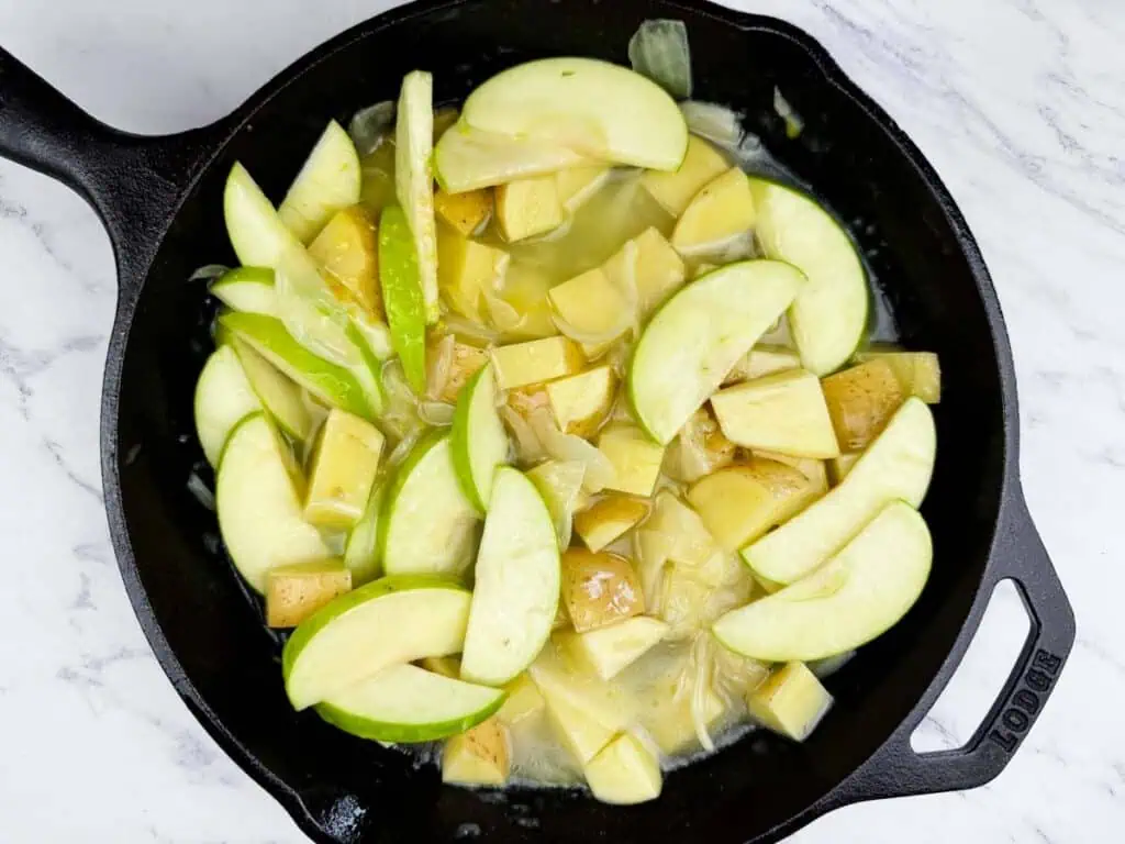 A cast iron skillet containing sliced green apples and chopped potatoes cooking in a light sauce on a marble countertop.