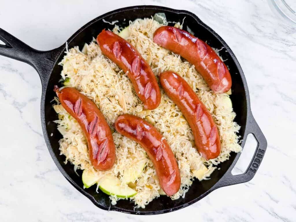 A cast iron skillet containing sliced sausage links arranged on top of a layer of sauerkraut and chopped vegetables.