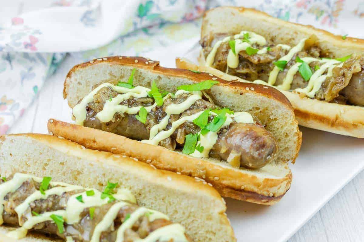 Three Beer Braised Brats in toasted buns topped with saut&eacute;ed onions, chopped parsley, and a drizzle of yellow sauce, served on a white plate.