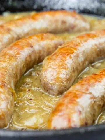 Five Beer Braised Bratwurst cooking in a skillet with a thick, light brown gravy or sauce.