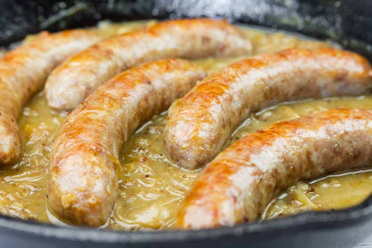 Five Beer Braised Bratwurst cooking in a skillet with a thick, light brown gravy or sauce.