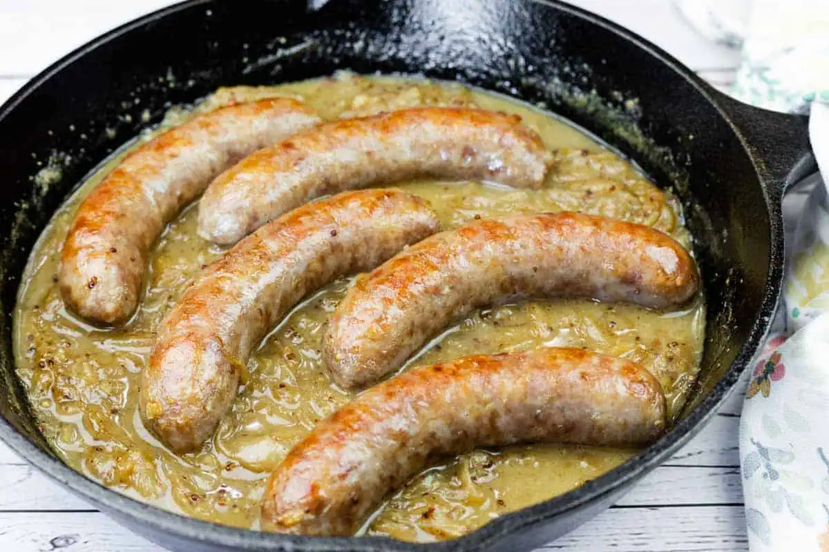 Five browned sausages simmer in onion gravy inside a black cast iron skillet on a light wooden surface.