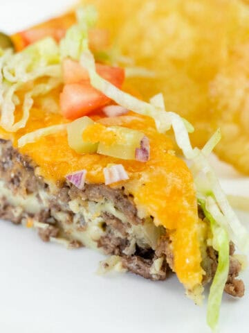 A slice of cheeseburger pie topped with shredded lettuce, diced tomato, onions, and pickles, served with potato chips on a white plate.