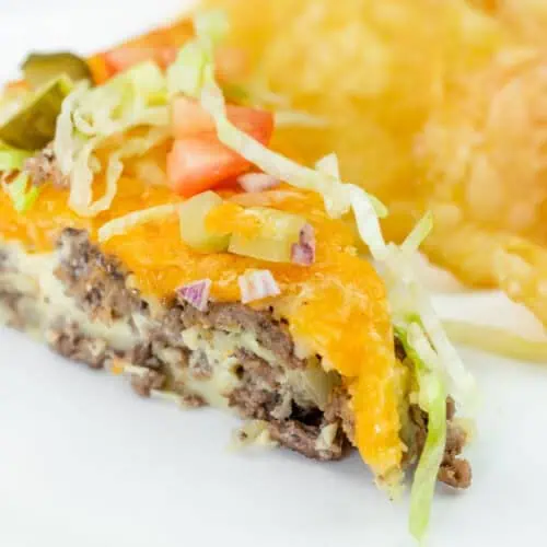 A slice of cheeseburger pie topped with shredded lettuce, diced tomato, onions, and pickles, served with potato chips on a white plate.