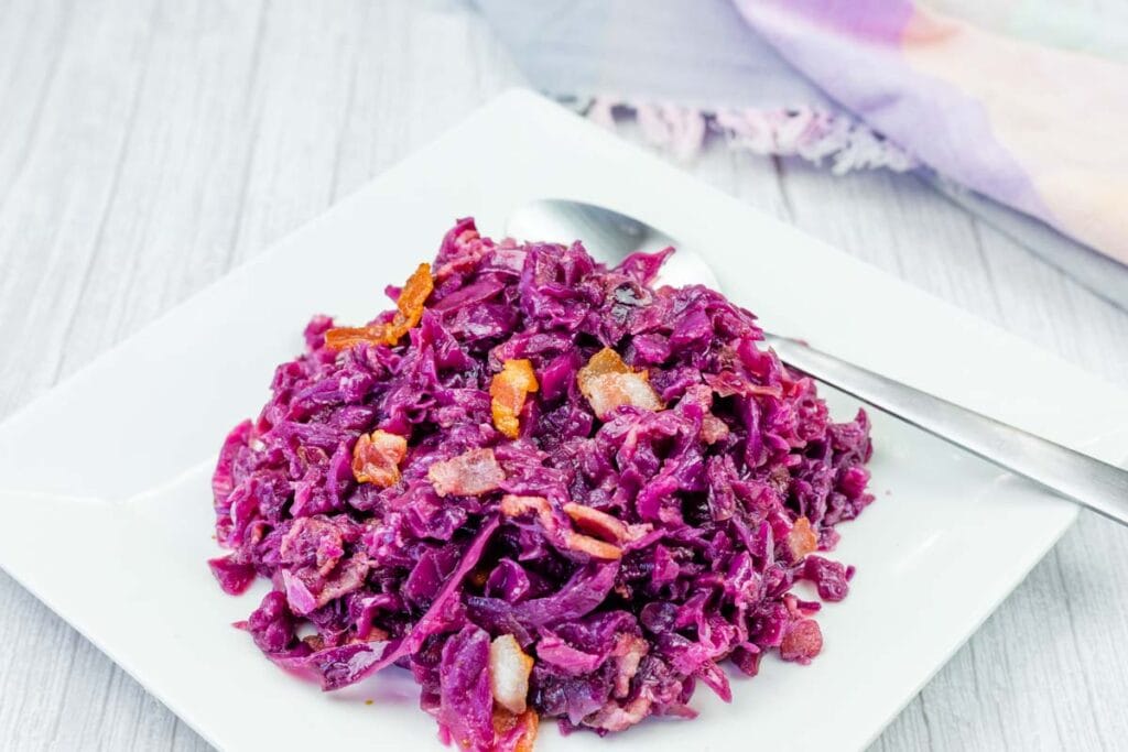 A serving of cooked red cabbage with pieces of bacon on a white square plate, accompanied by a spoon.