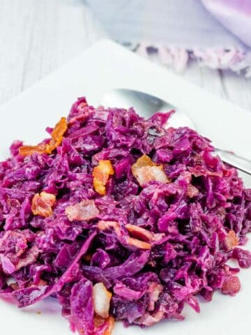 A serving of cooked red cabbage with pieces of bacon on a white square plate, accompanied by a spoon.