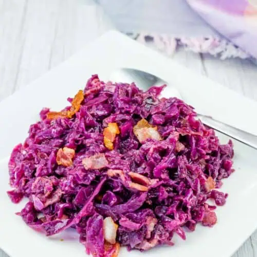 A serving of cooked red cabbage with pieces of bacon on a white square plate, accompanied by a spoon.