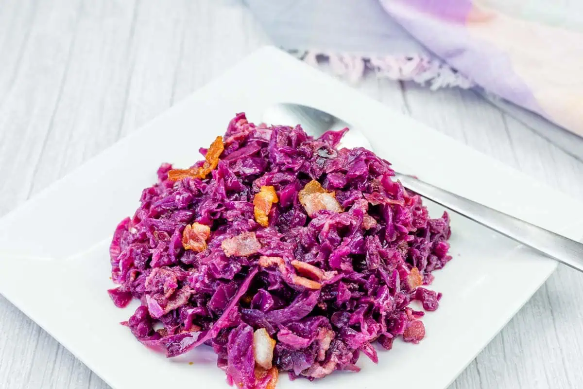 A serving of cooked red cabbage with pieces of bacon and carrots on a white square plate, with a spoon beside the food.