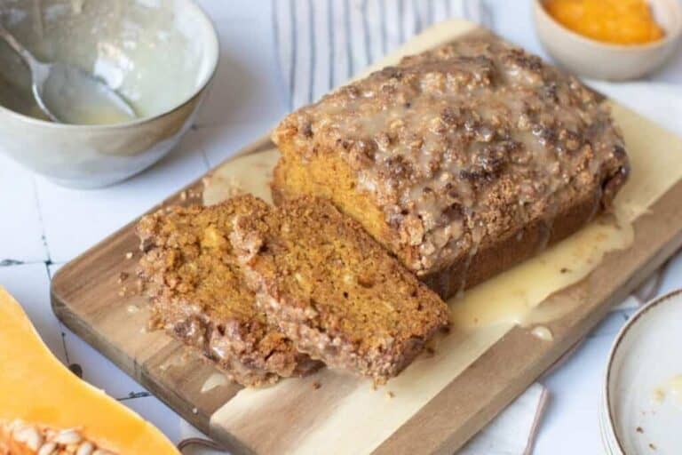 A loaf of glazed bread with two slices cut, displayed on a wooden board, surrounded by bowls and a butternut squash.