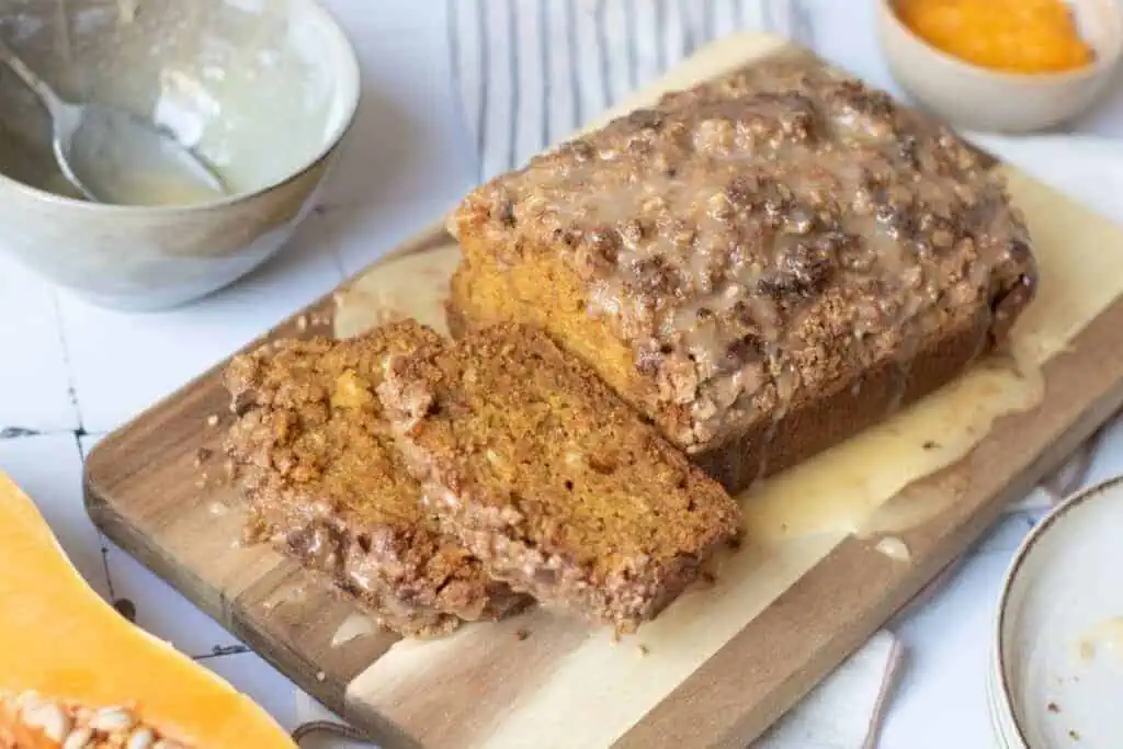 A loaf of glazed pumpkin bread with two slices cut sits on a wooden cutting board, surrounded by bowls containing glaze and orange puree.